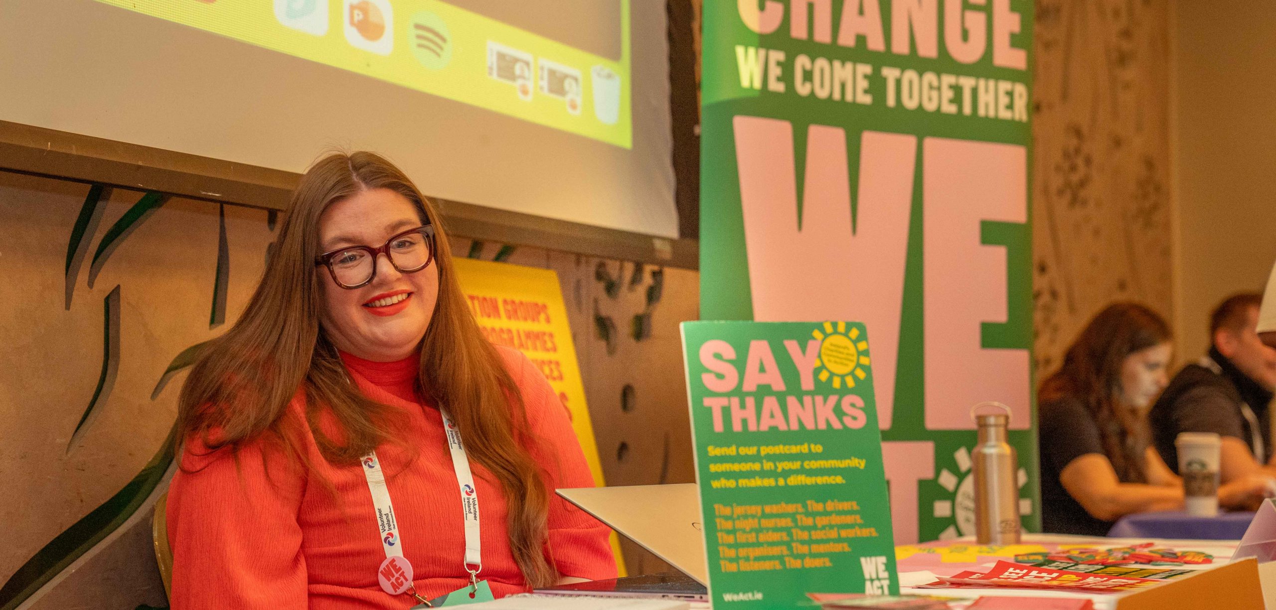 A smiling woman with glasses and long hair sits at a table in front of several signs